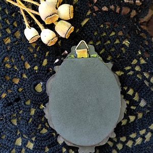 May include: An oval wall hanging with a dark grey surface and an ornate frame. A metal hanger is attached to the back. Dried poppy seed heads are arranged near the top, set against a black, open-weave fabric.