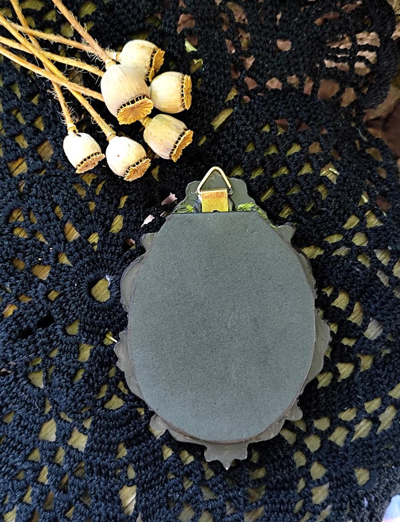 May include: An oval wall hanging with a dark grey surface and an ornate frame. A metal hanger is attached to the back. Dried poppy seed heads are arranged near the top, set against a black, open-weave fabric.
