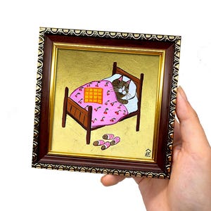Puede incluir: Una pintura enmarcada de un gato durmiendo en una cama con una manta rosa con estampado de cerezas. El gato es marr&oacute;n y blanco con una manta amarilla encima. La cama es de madera marr&oacute;n con un fondo dorado. Hay dos pares de zapatillas rosas y blancas en el suelo.