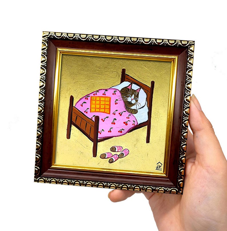 Puede incluir: Una pintura enmarcada de un gato durmiendo en una cama con una manta rosa con estampado de cerezas. El gato es marr&oacute;n y blanco con una manta amarilla encima. La cama es de madera marr&oacute;n con un fondo dorado. Hay dos pares de zapatillas rosas y blancas en el suelo.
