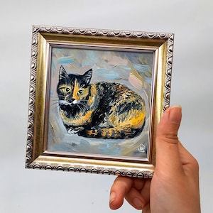 Pode incluir: Uma pintura a &oacute;leo de um gato tartaruga com pelo preto e laranja e olhos amarelos, feita com pinceladas vis&iacute;veis sobre um fundo cinza claro. A obra de arte est&aacute; inserida numa moldura quadrada de tom dourado com bordas ornamentadas.