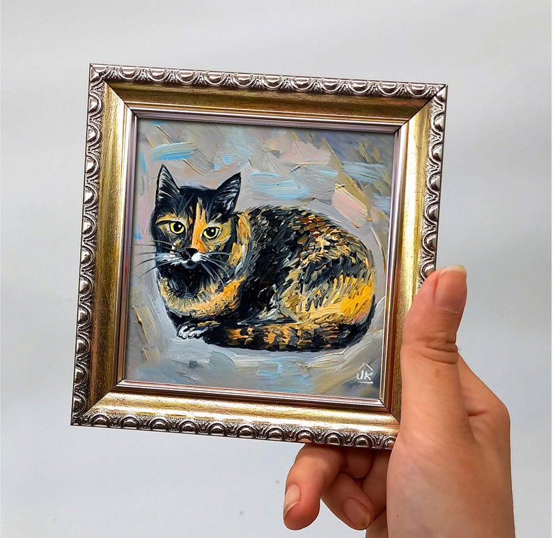 Pode incluir: Uma pintura a &oacute;leo de um gato tartaruga com pelo preto e laranja e olhos amarelos, feita com pinceladas vis&iacute;veis sobre um fundo cinza claro. A obra de arte est&aacute; inserida numa moldura quadrada de tom dourado com bordas ornamentadas.