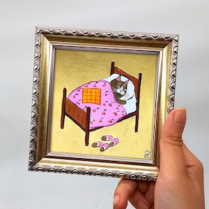 Puede incluir: Una pintura enmarcada de un gato durmiendo en una cama con una manta rosa con estampado de cerezas y una almohada a cuadros amarillos. La cama tiene un marco de madera y hay zapatillas rosas y blancas en el suelo. La pintura est&aacute; firmada "JK" en la esquina inferior derecha.