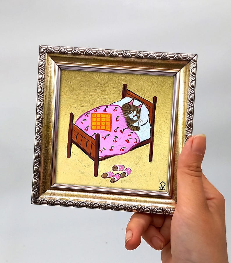Puede incluir: Una pintura enmarcada de un gato durmiendo en una cama con una manta rosa con estampado de cerezas y una almohada a cuadros amarillos. La cama tiene un marco de madera y hay zapatillas rosas y blancas en el suelo. La pintura est&aacute; firmada "JK" en la esquina inferior derecha.