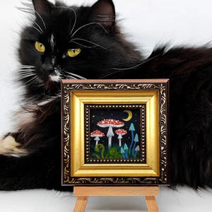 May include: A framed painting depicting a whimsical forest scene with red and white mushrooms, blue mushrooms, and a crescent moon. The painting is on a small easel.