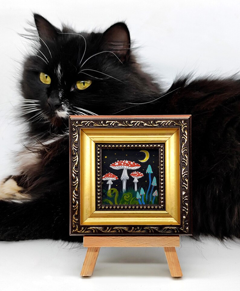 May include: A framed painting depicting a whimsical forest scene with red and white mushrooms, blue mushrooms, and a crescent moon. The painting is on a small easel.