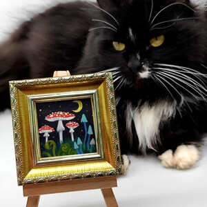May include: A small framed painting depicting a whimsical forest scene with red and white mushrooms, blue flowers, and a crescent moon. The painting is on a wooden easel with a black and white cat sitting in the background.