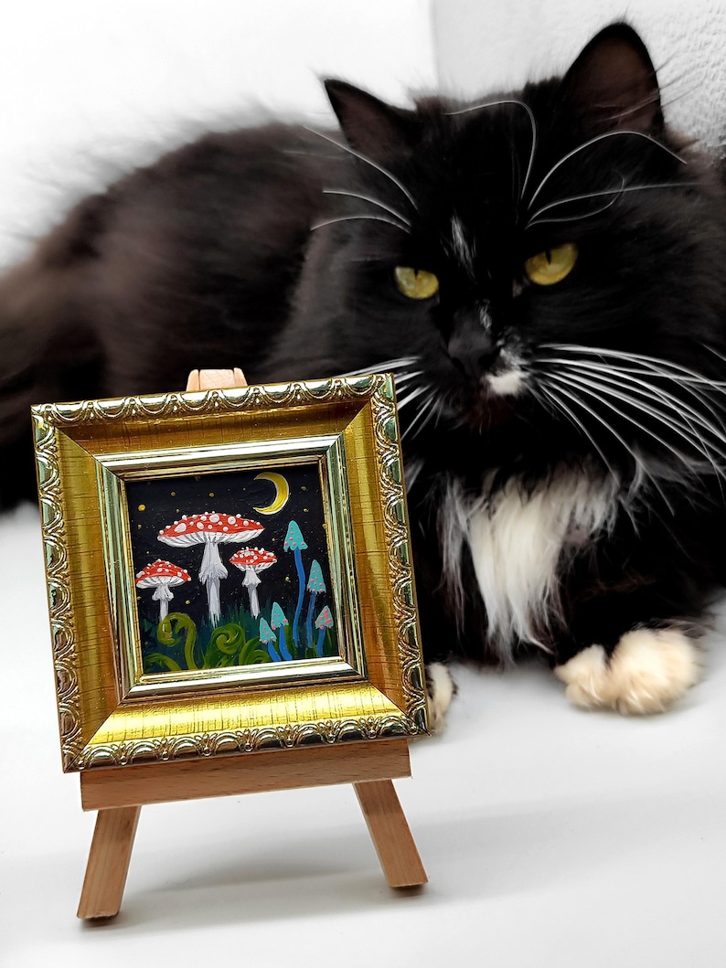 May include: A small framed painting depicting a whimsical forest scene with red and white mushrooms, blue flowers, and a crescent moon. The painting is on a wooden easel with a black and white cat sitting in the background.