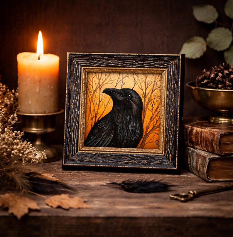 Pode incluir: Uma obra de arte emoldurada com um corvo preto contra um fundo laranja e amarelo com &aacute;rvores nuas. A obra de arte est&aacute; em uma moldura de madeira escura. Uma vela acesa, livros antigos e itens decorativos s&atilde;o dispostos ao redor da obra de arte.