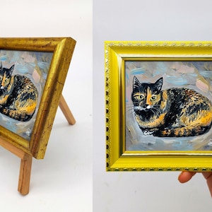 Pode incluir: Uma pequena pintura quadrada de um gato tartaruga com pelo preto, laranja e branco, feita com pinceladas vis&iacute;veis. A obra est&aacute; emoldurada numa moldura amarela brilhante e ornamentada com uma borda texturizada sobre um cavalete de madeira.
