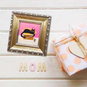 May include: A framed painting of a black cat sitting in a cardboard box with the word "FRAGILE" written on it. The box is on a pink background. The frame is gold and has a decorative border. The word "MOM" is spelled out in wooden letters below the frame.