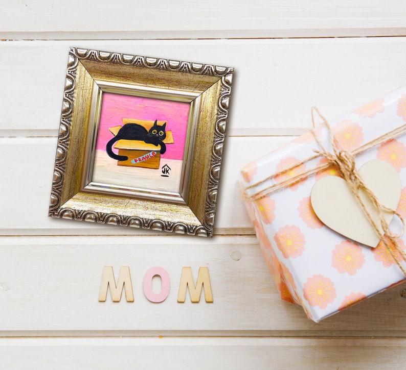 May include: A framed painting of a black cat sitting in a cardboard box with the word "FRAGILE" written on it. The box is on a pink background. The frame is gold and has a decorative border. The word "MOM" is spelled out in wooden letters below the frame.