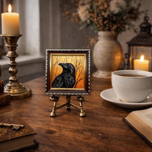 Pode incluir: Uma pequena obra de arte emoldurada de um corvo preto contra um fundo laranja e dourado, exibida em um cavalete de lat&atilde;o em miniatura. A cena inclui uma vela acesa, livros e uma x&iacute;cara de ch&aacute; em uma mesa de madeira.