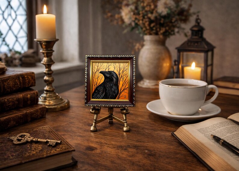 Pode incluir: Uma pequena obra de arte emoldurada de um corvo preto contra um fundo laranja e dourado, exibida em um cavalete de lat&atilde;o em miniatura. A cena inclui uma vela acesa, livros e uma x&iacute;cara de ch&aacute; em uma mesa de madeira.