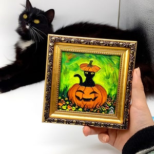 Op de afbeelding: Een vierkant olieverfschilderij in een sierlijke gouden lijst met een zwarte kat die in een uitgesneden oranje pompoen zit met een pompoendeksel op zijn kop. De achtergrond is levendig groen met gestructureerde penseelstreken en bladeren.