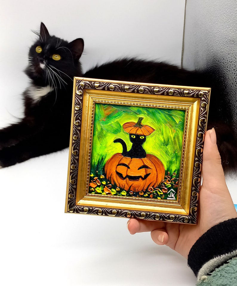 Op de afbeelding: Een vierkant olieverfschilderij in een sierlijke gouden lijst met een zwarte kat die in een uitgesneden oranje pompoen zit met een pompoendeksel op zijn kop. De achtergrond is levendig groen met gestructureerde penseelstreken en bladeren.
