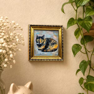 Pode incluir: Uma pintura a &oacute;leo emoldurada de formato quadrado apresentando um gato de pelagem tricolor preta, laranja e branca, retratado em posi&ccedil;&atilde;o de repouso enrolado contra um fundo texturizado azul-claro e cinza, em uma moldura dourada ornamentada.