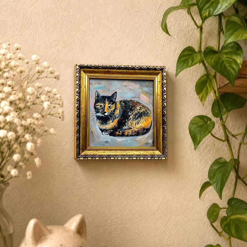 Pode incluir: Uma pintura a &oacute;leo emoldurada de formato quadrado apresentando um gato de pelagem tricolor preta, laranja e branca, retratado em posi&ccedil;&atilde;o de repouso enrolado contra um fundo texturizado azul-claro e cinza, em uma moldura dourada ornamentada.