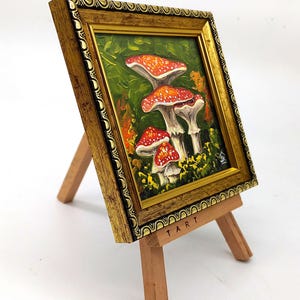 May include: A framed painting of red and white mushrooms in a green forest setting. The painting is on a small wooden easel with the word "TART" printed on the base.