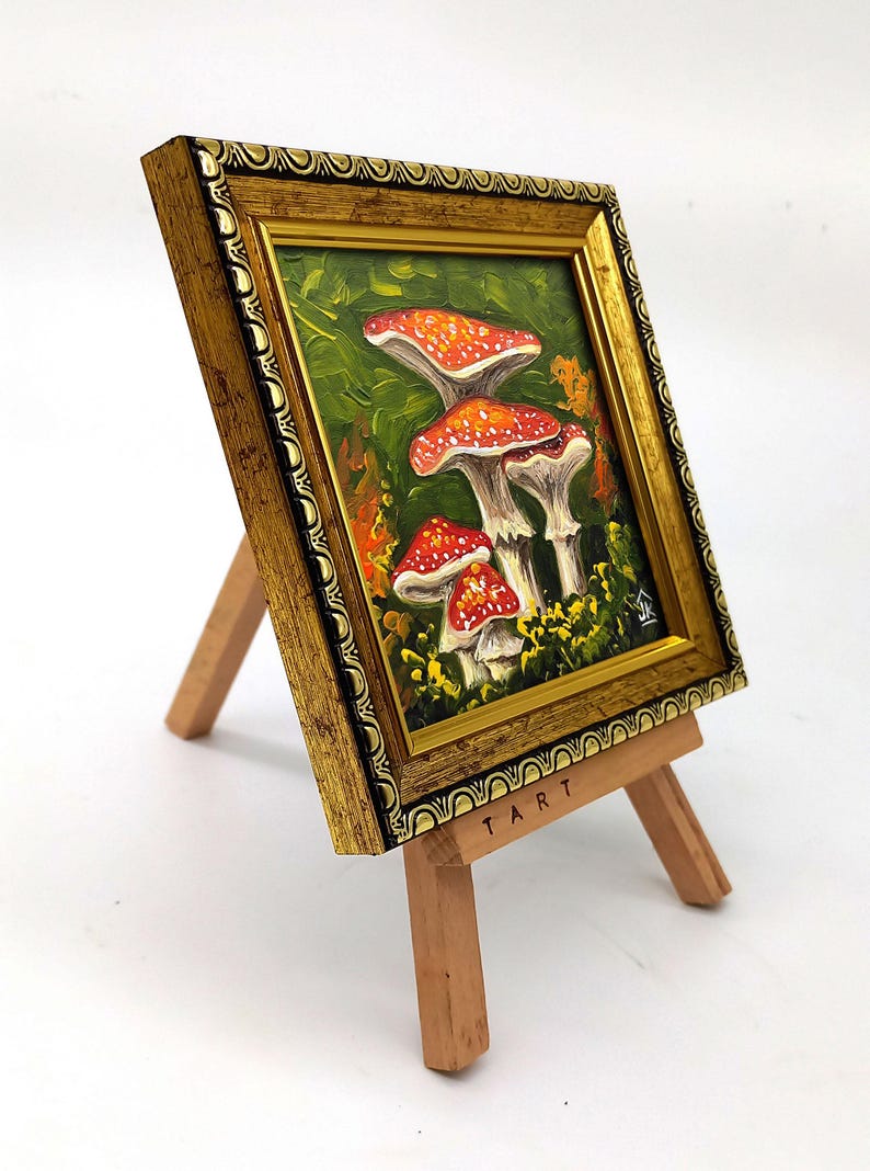 May include: A framed painting of red and white mushrooms in a green forest setting. The painting is on a small wooden easel with the word "TART" printed on the base.