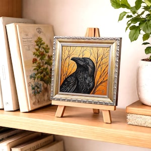 Pode incluir: Uma obra de arte emoldurada com um corvo preto contra um fundo de &aacute;rvores nuas e um c&eacute;u laranja. A obra de arte &eacute; exibida em um pequeno cavalete de madeira. A moldura tem um acabamento dourado e prateado. A obra de arte est&aacute; em uma prateleira de madeira com livros e uma planta em vaso.