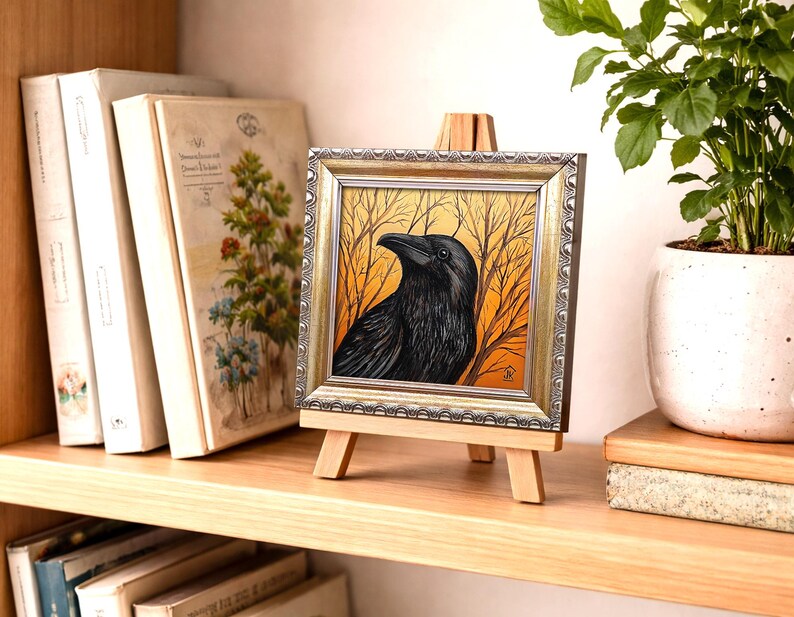 Pode incluir: Uma obra de arte emoldurada com um corvo preto contra um fundo de &aacute;rvores nuas e um c&eacute;u laranja. A obra de arte &eacute; exibida em um pequeno cavalete de madeira. A moldura tem um acabamento dourado e prateado. A obra de arte est&aacute; em uma prateleira de madeira com livros e uma planta em vaso.