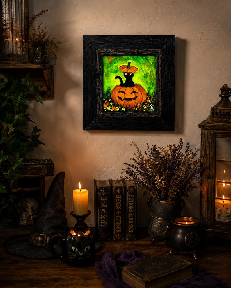 Op de afbeelding: Een vierkant ingelijst schilderij met een zwarte kat die in een uitgesneden oranje pompoen zit, met een kleiner pompoenstuk op de kop, tegen een levendige groene achtergrond met gevallen herfstbladeren aan de onderkant.