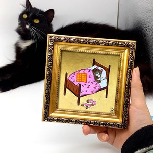 Puede incluir: Una pintura enmarcada de un gato durmiendo en una cama rosa con estampado de cerezas. El gato es marr&oacute;n y blanco y tiene los ojos cerrados. La cama est&aacute; sobre un fondo dorado. La pintura est&aacute; en un marco dorado con un borde marr&oacute;n oscuro.
