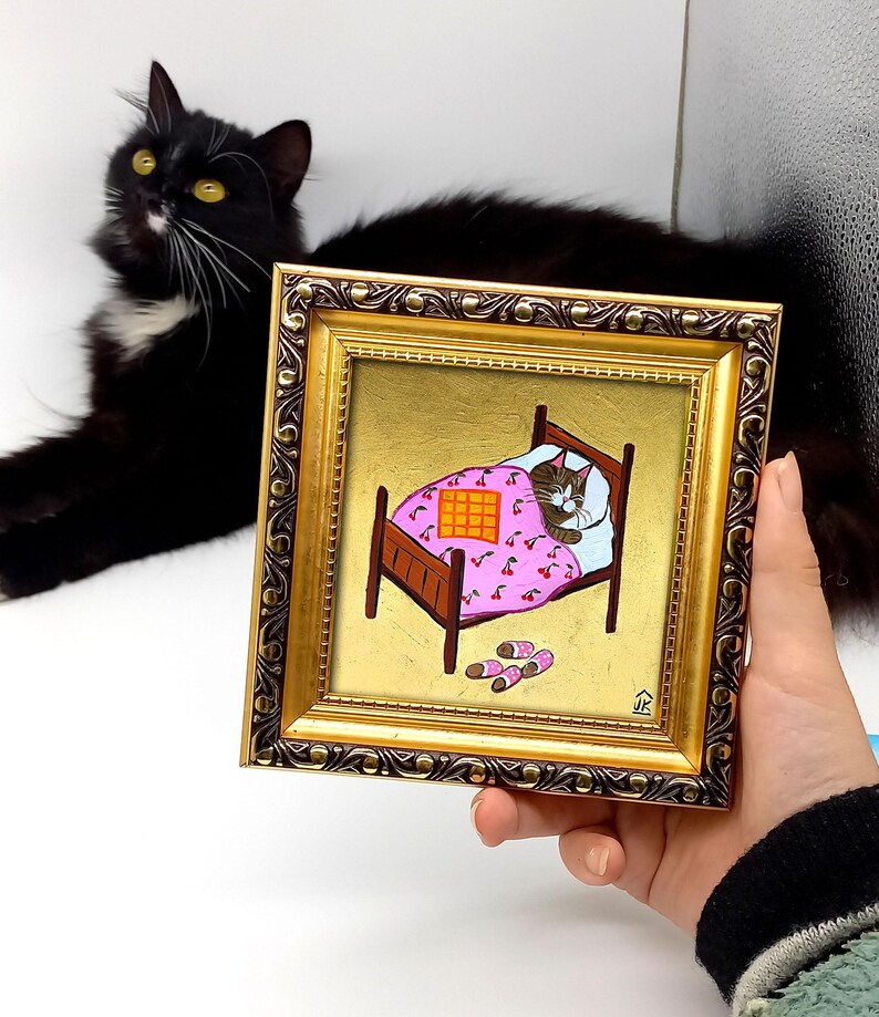 Puede incluir: Una pintura enmarcada de un gato durmiendo en una cama rosa con estampado de cerezas. El gato es marr&oacute;n y blanco y tiene los ojos cerrados. La cama est&aacute; sobre un fondo dorado. La pintura est&aacute; en un marco dorado con un borde marr&oacute;n oscuro.