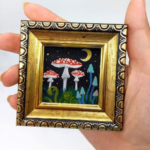 May include: A small framed painting of red and white mushrooms with blue and green accents on a black background. The painting depicts a moon and stars in the night sky.