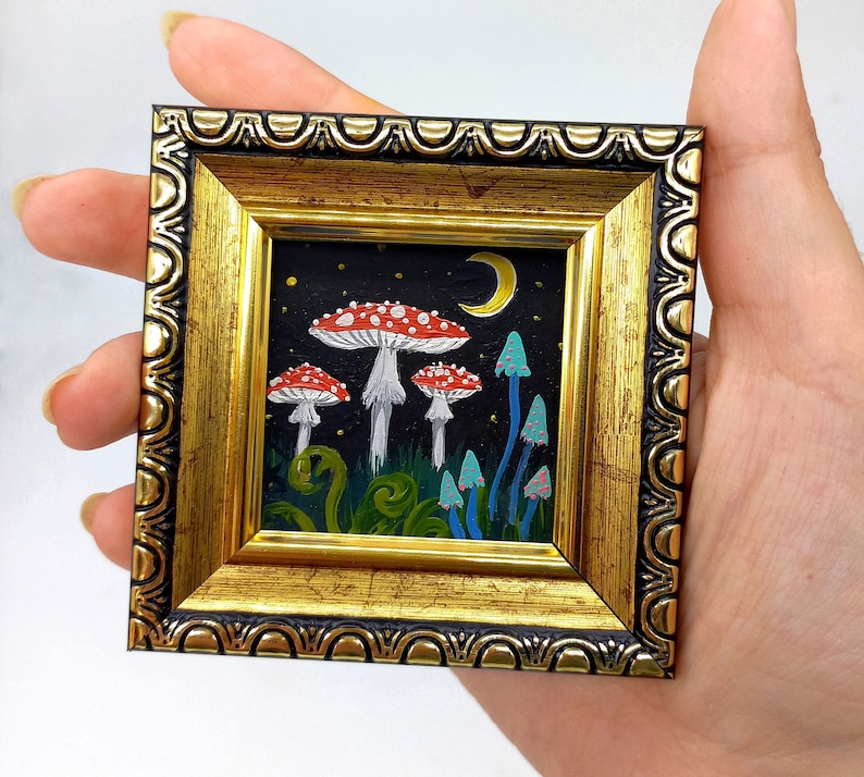 May include: A small framed painting of red and white mushrooms with blue and green accents on a black background. The painting depicts a moon and stars in the night sky.