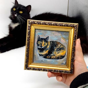 Pode incluir: Uma pequena pintura a &oacute;leo quadrada de um gato tartaruga numa moldura dourada decorativa. A obra apresenta pinceladas vis&iacute;veis e um fundo azul claro. O gato est&aacute; representado numa posi&ccedil;&atilde;o de repouso com olhos amarelos.