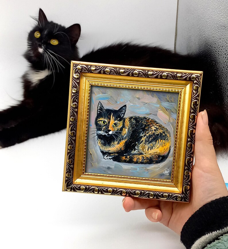 Pode incluir: Uma pequena pintura a &oacute;leo quadrada de um gato tartaruga numa moldura dourada decorativa. A obra apresenta pinceladas vis&iacute;veis e um fundo azul claro. O gato est&aacute; representado numa posi&ccedil;&atilde;o de repouso com olhos amarelos.