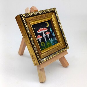 May include: A small framed painting depicting a whimsical forest scene with red and white mushrooms, blue flowers, and a crescent moon in the night sky.