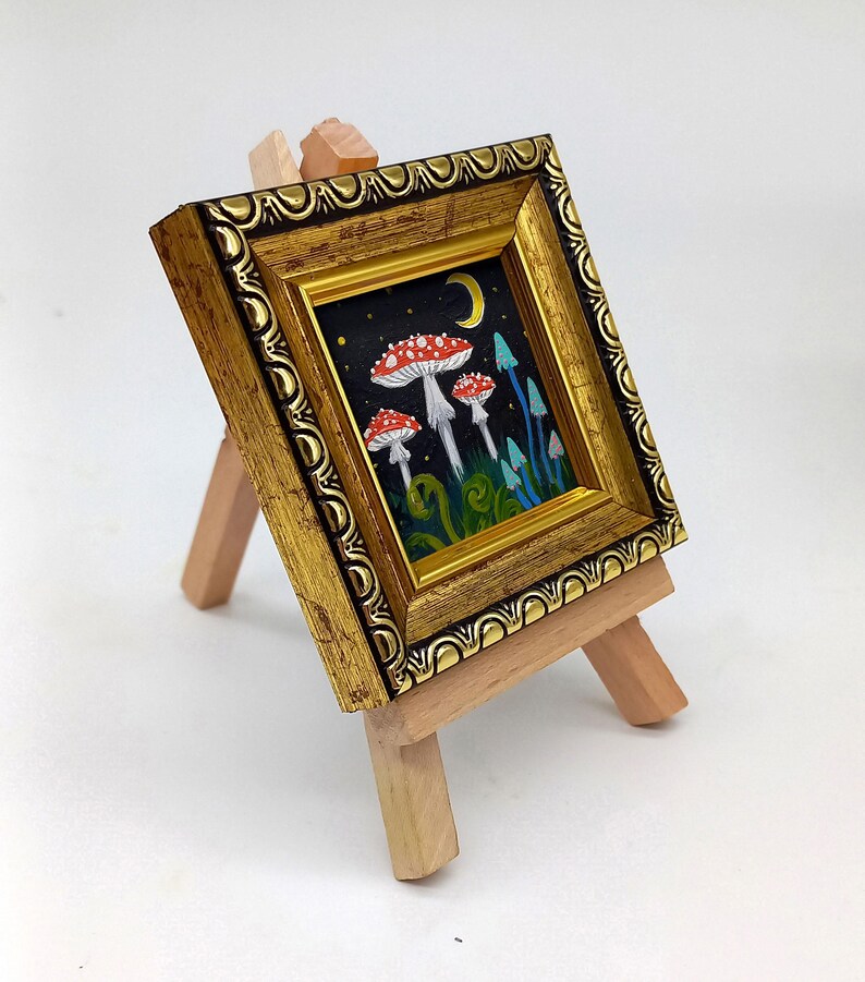 May include: A small framed painting depicting a whimsical forest scene with red and white mushrooms, blue flowers, and a crescent moon in the night sky.