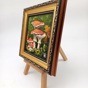 May include: A framed painting of three red and white mushrooms with white spots on a green background. The painting is on a small wooden easel.