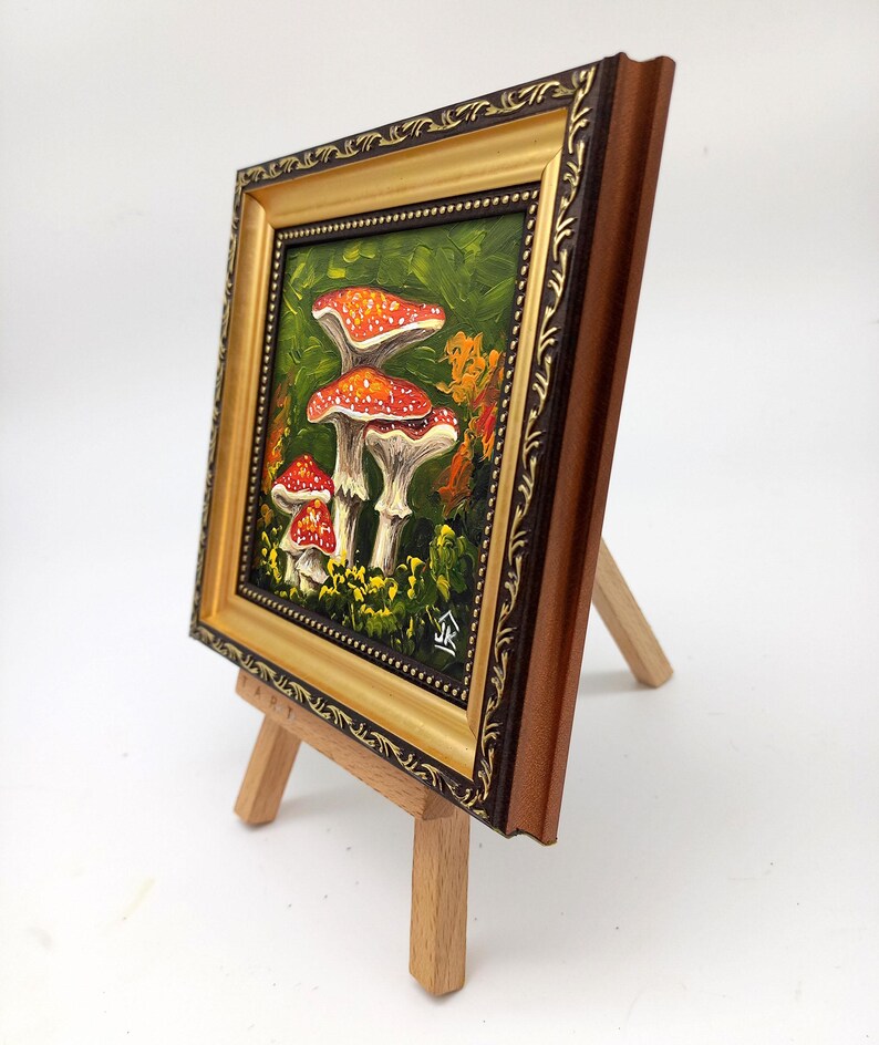 May include: A framed painting of three red and white mushrooms with white spots on a green background. The painting is on a small wooden easel.