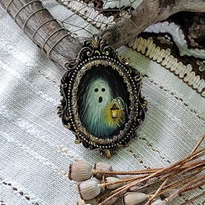 May include: An oval-shaped, black and gold framed painting of a ghost holding a lantern. The ghost is white with a dark background. The frame has ornate detailing. The painting is on a textured, off-white surface.