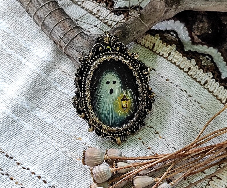 May include: An oval-shaped, black and gold framed painting of a ghost holding a lantern. The ghost is white with a dark background. The frame has ornate detailing. The painting is on a textured, off-white surface.