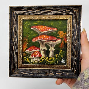 May include: A framed painting of red and white mushrooms with white spots in a green forest setting. The painting is signed "JK" in the bottom right corner.