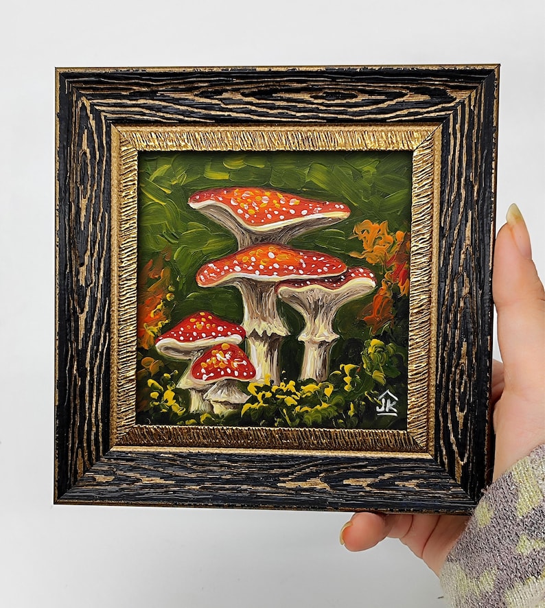 May include: A framed painting of red and white mushrooms with white spots in a green forest setting. The painting is signed "JK" in the bottom right corner.