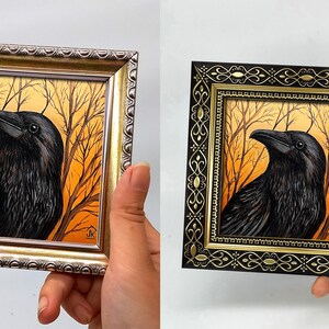 Pode incluir: Duas obras de arte emolduradas de um corvo. Uma moldura dourada, a outra preta com detalhes dourados. O corvo preto com penas detalhadas, contra um fundo laranja e castanho de &aacute;rvores nuas. A obra de arte &eacute; segurada por uma m&atilde;o.