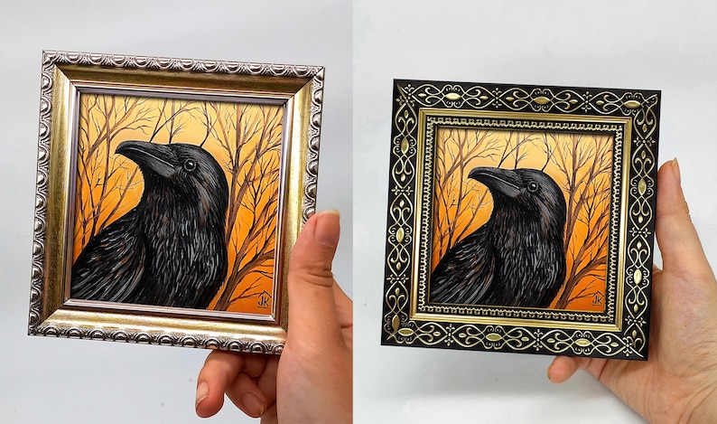 Pode incluir: Duas obras de arte emolduradas de um corvo. Uma moldura dourada, a outra preta com detalhes dourados. O corvo preto com penas detalhadas, contra um fundo laranja e castanho de &aacute;rvores nuas. A obra de arte &eacute; segurada por uma m&atilde;o.