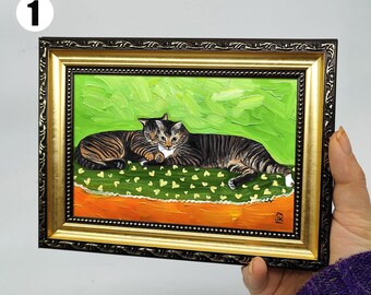 May include: A framed painting of two tabby cats resting together. The artwork features a green background with heart-shaped details, an orange base, and a gold and black ornate frame. The cats are brown and black with white markings.