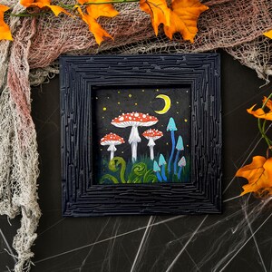 May include: A framed painting depicting a whimsical forest scene with red and white mushrooms, blue and green plants, and a crescent moon in the night sky.