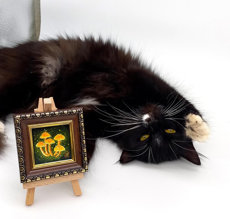 May include: A small framed painting of a cluster of yellow mushrooms on a green background. The painting is on a wooden easel and a black and white cat is lying on its back in front of it.