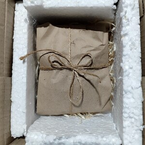 May include: A parcel wrapped in brown paper and secured with twine, placed within a cardboard box lined with white foam padding. The packaging suggests a focus on presentation and protection during transit. The overall aesthetic is rustic and eco-conscious.