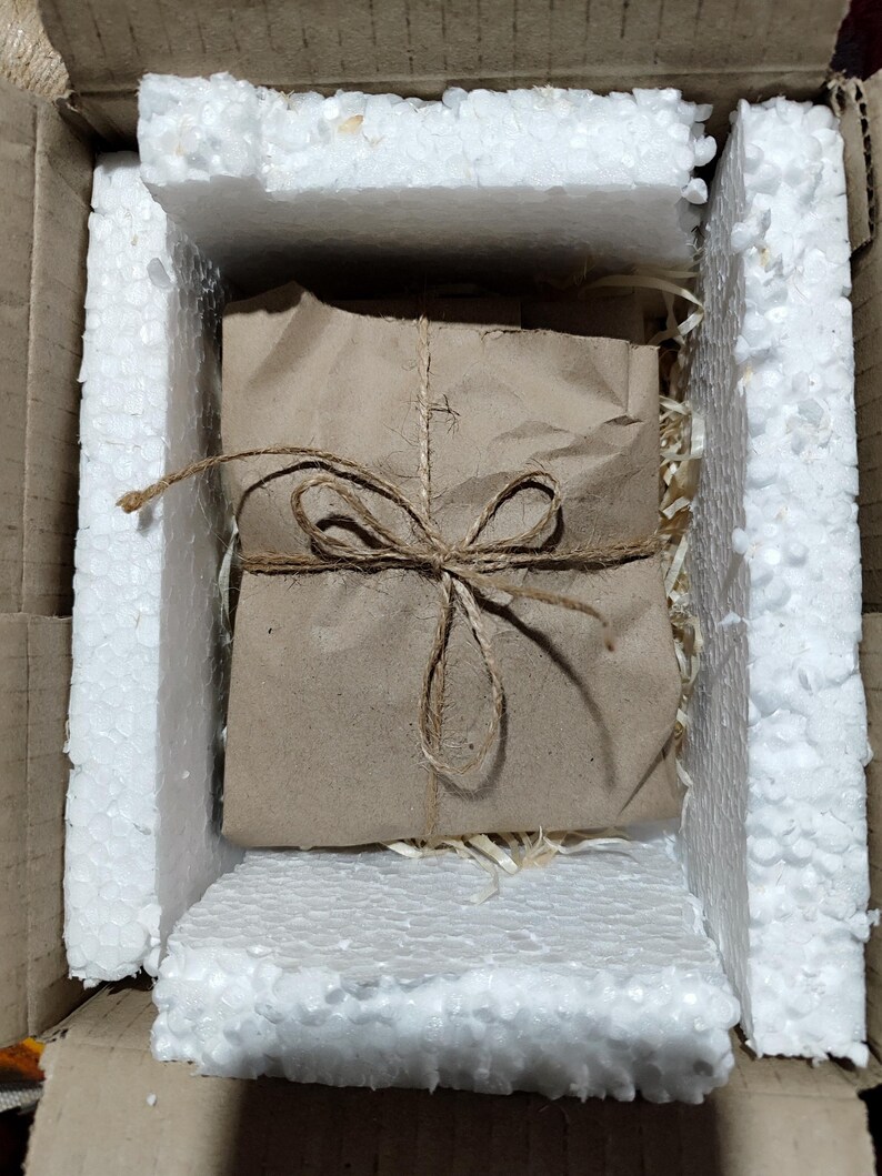 May include: A parcel wrapped in brown paper and secured with twine, placed within a cardboard box lined with white foam padding. The packaging suggests a focus on presentation and protection during transit. The overall aesthetic is rustic and eco-conscious.
