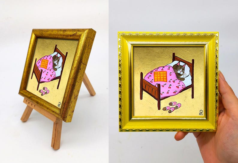 Puede incluir: Una pintura enmarcada de un gato durmiendo en una cama con una manta de estampado floral rosa y blanco. El gato es marr&oacute;n y blanco y tiene los ojos cerrados. La cama es de madera y tiene una manta a cuadros encima de la manta floral. Hay dos pares de zapatillas en el suelo al lado de la cama.