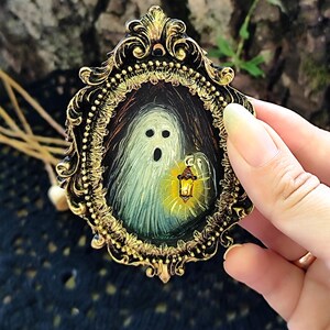May include: A miniature painting of a ghost with a lantern, set within an ornate gold and black frame. The ghost is depicted with a surprised expression. The frame is held by a hand, showcasing the intricate details of the artwork.